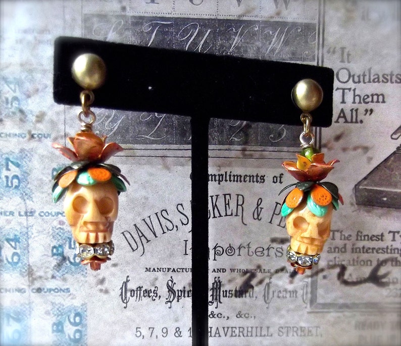 May include: A pair of skull-shaped earrings with orange, turquoise, and white enamel details. The earrings are hanging from a black display stand. The text "Outlasts Them All." is visible in the background.