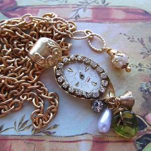 May include: A gold chain necklace with a vintage watch pendant, adorned with rhinestones. The watch face is white with black Roman numeral markings. The necklace also features a variety of charms, including a pearl, a leaf, and a small bell.