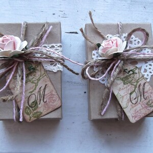 May include: Two small brown gift boxes tied with twine and pink ribbon. Each box has a white lace doily, a pink flower, and a tag with the letter "M" on it.