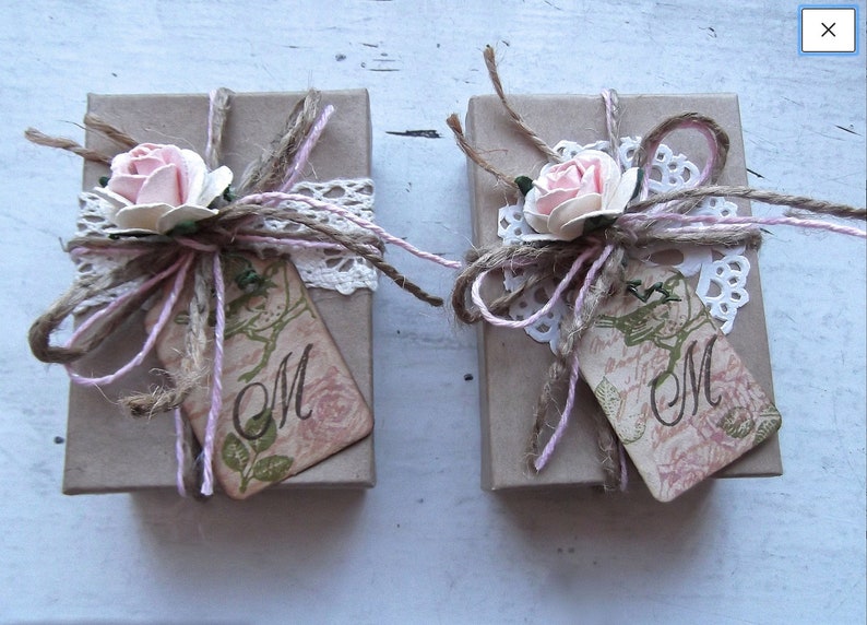 May include: Two small brown gift boxes tied with twine and pink ribbon. Each box has a white lace doily, a pink flower, and a tag with the letter "M" on it.