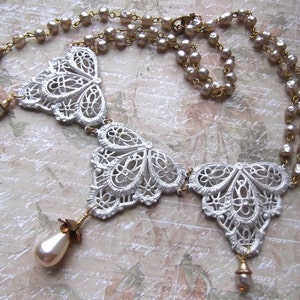 May include: A white, ornate necklace with three butterfly-shaped pendants and a pearl drop on each end. The necklace is made of metal and has a delicate, lace-like design.