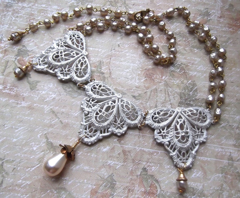 May include: A white, ornate necklace with three butterfly-shaped pendants and a pearl drop on each end. The necklace is made of metal and has a delicate, lace-like design.