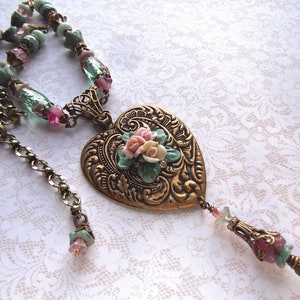 Puede incluir: Collar de estilo vintage con un colgante en forma de coraz&oacute;n dorado. El colgante presenta un patr&oacute;n en relieve y un dise&ntilde;o floral con flores rosas, blancas y verdes. La cadena tiene cuentas rosas y verdes.