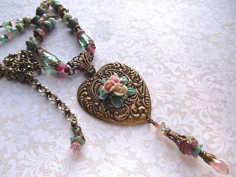 Puede incluir: Collar de estilo vintage con un colgante en forma de coraz&oacute;n dorado. El colgante presenta un patr&oacute;n en relieve y un dise&ntilde;o floral con flores rosas, blancas y verdes. La cadena tiene cuentas rosas y verdes.