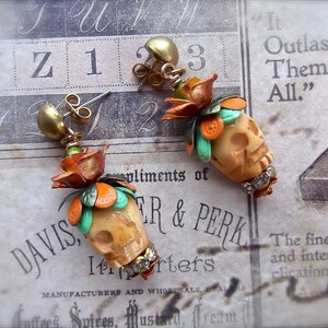 May include: A pair of dangle earrings with a skull charm, orange and green floral accents, and rhinestones. The earrings are on a white and brown patterned surface with the text "It Outlasts Them All." and "DAVIS, TR & PERK"