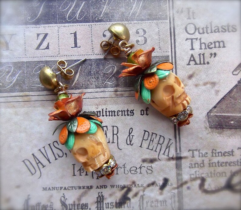 May include: A pair of dangle earrings with a skull charm, orange and green floral accents, and rhinestones. The earrings are on a white and brown patterned surface with the text "It Outlasts Them All." and "DAVIS, TR & PERK"