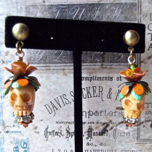 May include: A pair of skull-shaped earrings with orange and green floral accents. The earrings are made of white ceramic and have rhinestones on the bottom.