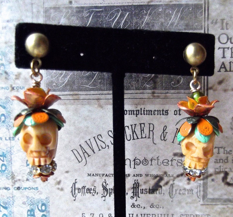 May include: A pair of skull-shaped earrings with orange and green floral accents. The earrings are made of white ceramic and have rhinestones on the bottom.