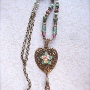 Puede incluir: Collar de estilo vintage con un colgante en forma de coraz&oacute;n en tono bronce, con detalles florales en relieve y un racimo de peque&ntilde;as rosas. La cadena incluye cuentas rosas, verdes y transparentes, y una borla decorativa. El dise&ntilde;o es rom&aacute;ntico y detallado.