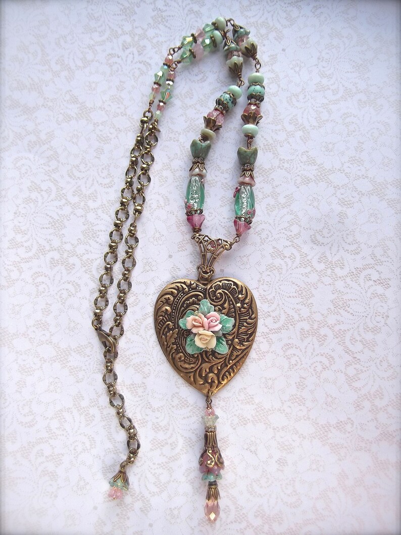 Puede incluir: Collar de estilo vintage con un colgante en forma de coraz&oacute;n en tono bronce, con detalles florales en relieve y un racimo de peque&ntilde;as rosas. La cadena incluye cuentas rosas, verdes y transparentes, y una borla decorativa. El dise&ntilde;o es rom&aacute;ntico y detallado.
