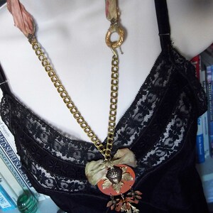 May include: A gold chain necklace with a large, orange, acorn-shaped pendant. The pendant is decorated with gold accents and a small, brown, fabric flower. The necklace is worn over a black lace top.