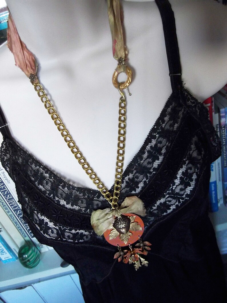 May include: A gold chain necklace with a large, orange, acorn-shaped pendant. The pendant is decorated with gold accents and a small, brown, fabric flower. The necklace is worn over a black lace top.
