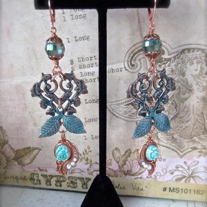 May include: A pair of copper and turquoise dangle earrings with a filigree design and a turquoise and white bead at the bottom. The earrings are hanging on a black stand.