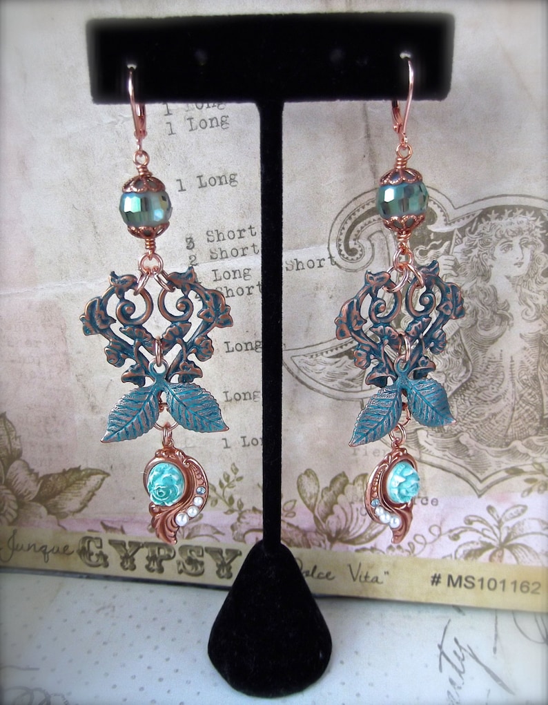 May include: A pair of copper and turquoise dangle earrings with a filigree design and a turquoise and white bead at the bottom. The earrings are hanging on a black stand.