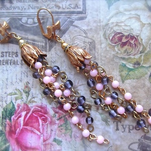 May include: A pair of gold tone earrings with pink and purple glass beads and a gold flower charm. The earrings are hanging from a gold chain.
