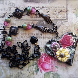 Pode incluir: Um colar de corrente preta com um pingente em forma de cora&ccedil;&atilde;o. O pingente &eacute; feito de cer&acirc;mica preta e branca e apresenta um design floral rosa e branco. O colar tamb&eacute;m inclui uma variedade de contas, incluindo pretas, brancas e verdes.