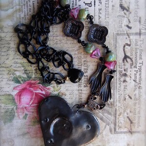 Pode incluir: Um colar de corrente preta com um pingente em forma de cora&ccedil;&atilde;o. O pingente &eacute; feito de metal e tem um acabamento desgastado. O colar &eacute; adornado com contas de vidro coloridas, pingentes de metal e duas m&atilde;os pretas.