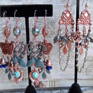 May include: A pair of copper and green statement earrings with a crown, bells, and leaves. The earrings are hanging from a black display stand.
