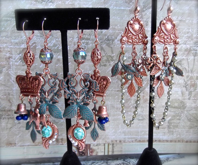 May include: A pair of copper and green statement earrings with a crown, bells, and leaves. The earrings are hanging from a black display stand.