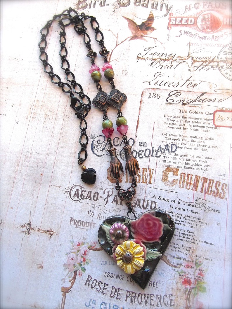Pode incluir: Um colar com um pingente em forma de cora&ccedil;&atilde;o preto com um design floral. O pingente &eacute; feito de cer&acirc;mica e apresenta flores rosa, amarelas e verdes. O colar &eacute; feito de corrente de metal preto com contas verdes e rosas.