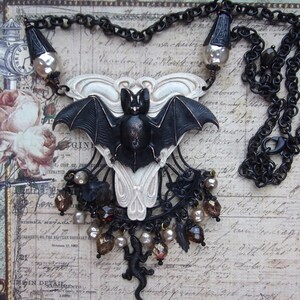 May include: A gothic-style necklace featuring a black bat pendant with outstretched wings, set against a white decorative backdrop. The necklace includes a black chain, decorative beads, and a small lizard charm. The background has vintage script.