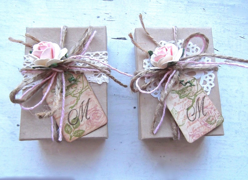 Puede incluir: Dos peque&ntilde;as cajas de regalo rectangulares envueltas en papel marr&oacute;n, adornadas con rosas rosas, hilo, encaje y etiquetas. Las etiquetas presentan la letra "M" y dise&ntilde;os florales, creando una est&eacute;tica r&uacute;stica y rom&aacute;ntica.