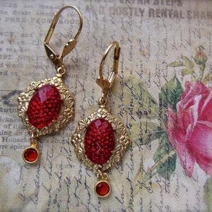 May include: A pair of gold earrings with red glass stones. The earrings are teardrop shaped and are accented with small gold beads.