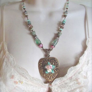 Puede incluir: Un collar de estilo vintage con un colgante en forma de coraz&oacute;n dorado adornado con flores. La cadena tiene cuentas rosas, verdes y azules. El colgante presenta patrones detallados y un dije colgante.