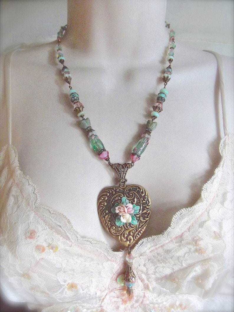 Puede incluir: Un collar de estilo vintage con un colgante en forma de coraz&oacute;n dorado adornado con flores. La cadena tiene cuentas rosas, verdes y azules. El colgante presenta patrones detallados y un dije colgante.