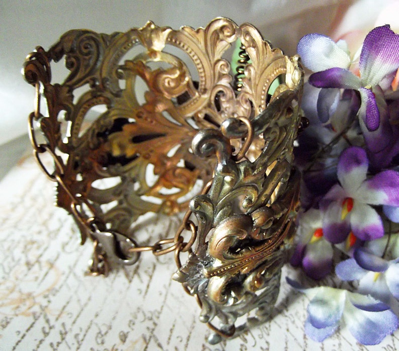 May include: A gold and silver ornate metal bracelet with a chain detail. The bracelet is intricately designed with floral patterns and has a vintage aesthetic.