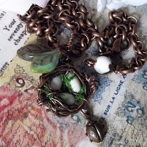 May include: A copper chain necklace with a bird's nest charm. The nest is made of wire and has three white eggs inside. The nest is decorated with green moss. The necklace also has a small, copper bird charm.