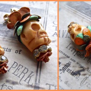 May include: A pair of skull-shaped earrings with orange, green, and white floral accents. The earrings are made of metal and have a vintage style.
