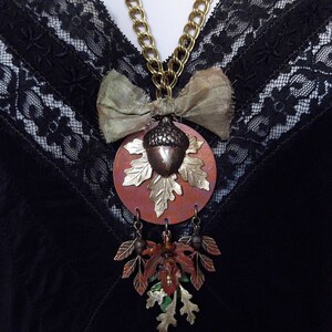May include: A gold chain necklace with a copper acorn pendant and a brown bow. The pendant is surrounded by a copper leaf design. The necklace is hanging on a black lace fabric.