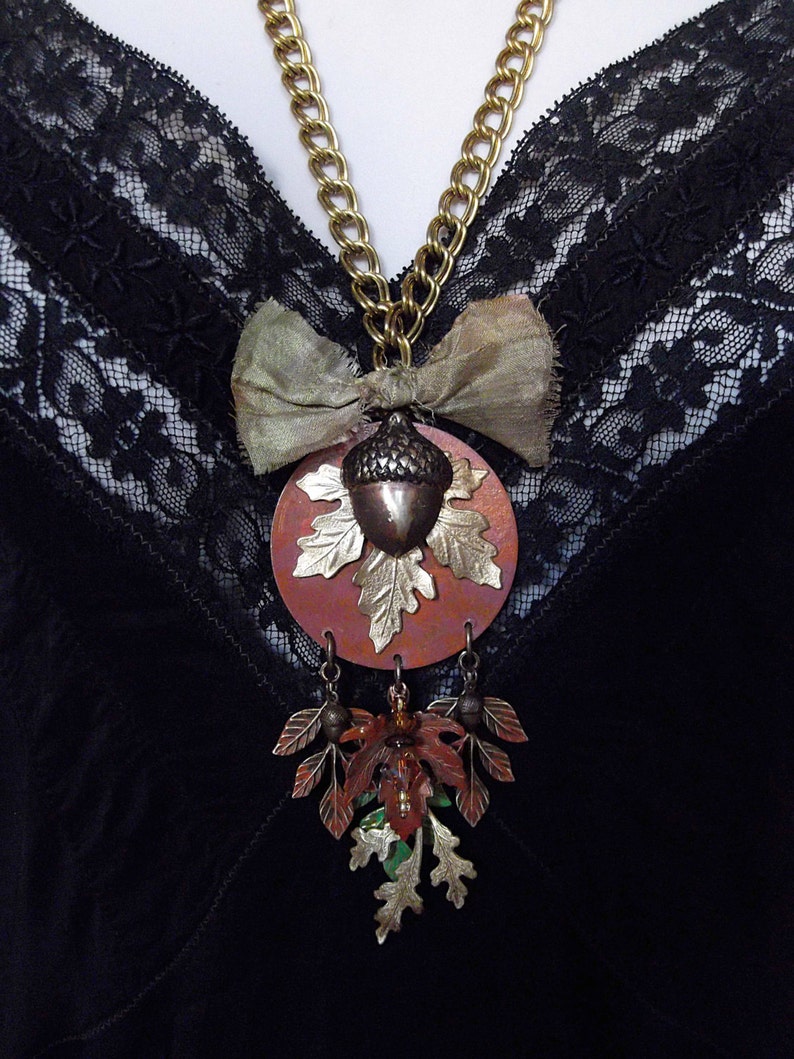 May include: A gold chain necklace with a copper acorn pendant and a brown bow. The pendant is surrounded by a copper leaf design. The necklace is hanging on a black lace fabric.