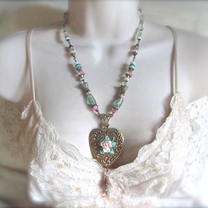 Puede incluir: Un collar de estilo vintage con un colgante en forma de coraz&oacute;n dorado adornado con un dise&ntilde;o floral. El collar presenta una cadena de cuentas con cuentas de colores pastel. El collar se muestra sobre un fondo blanco.