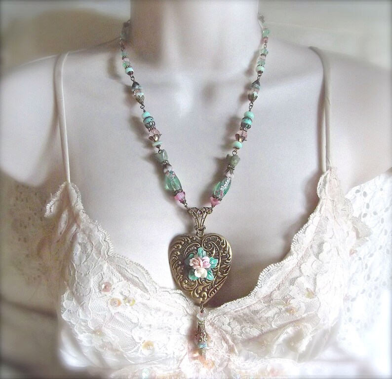 Puede incluir: Un collar de estilo vintage con un colgante en forma de coraz&oacute;n dorado adornado con un dise&ntilde;o floral. El collar presenta una cadena de cuentas con cuentas de colores pastel. El collar se muestra sobre un fondo blanco.