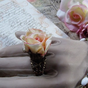 May include: A ring with a delicate, white and pink fabric rose, accented with a bronze-colored setting. The ring is displayed on a gloved hand with a white beaded bracelet.