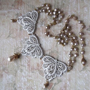 May include: A white filigree necklace with three butterfly-shaped pendants and a strand of pale pink pearls. The necklace has gold accents and is perfect for a vintage or bohemian style.