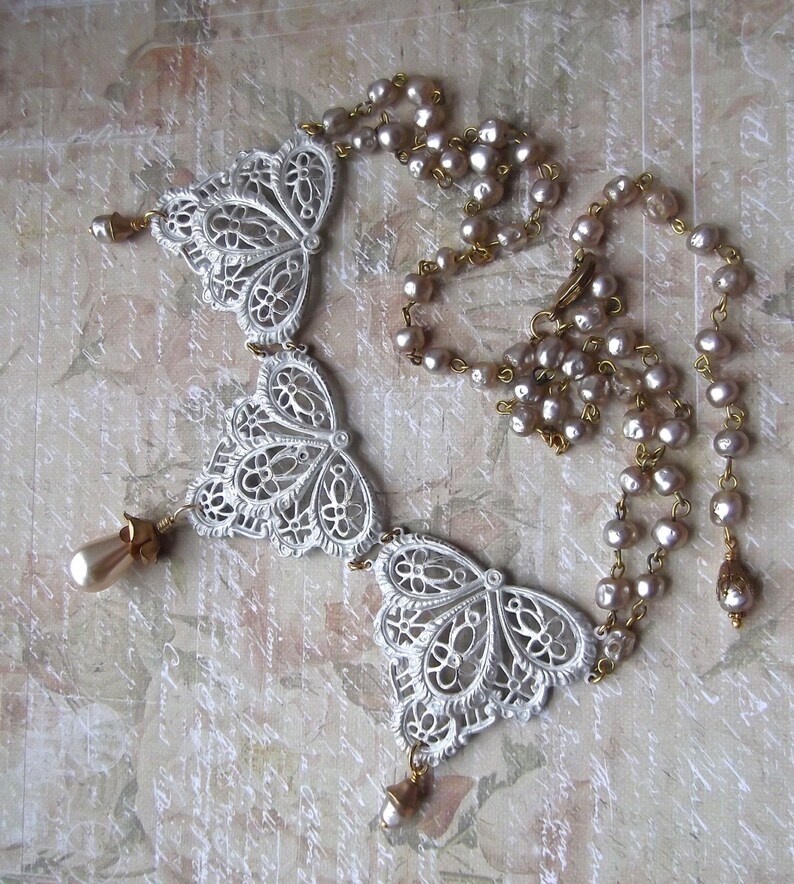 May include: A white filigree necklace with three butterfly-shaped pendants and a strand of pale pink pearls. The necklace has gold accents and is perfect for a vintage or bohemian style.