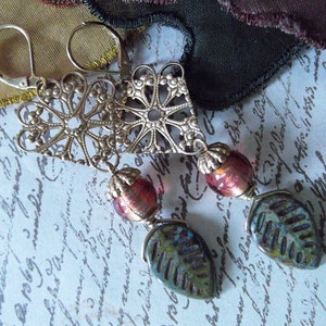 May include: Silver filigree drop earrings with lever-back fastenings. Each earring has a decorative metal top, a red and gold bead, and a leaf-shaped charm in blue and green. The earrings are shown against a pale blue background.