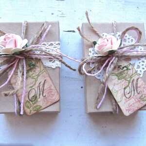 Pode incluir: Duas pequenas caixas de presente marrons amarradas com barbante e renda. Cada caixa tem uma flor rosa e uma etiqueta com a letra "M" nela.