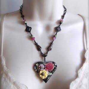 Pode incluir: Um colar de corrente preta com um pingente de cora&ccedil;&atilde;o de cer&acirc;mica com flores rosa, amarelas e verdes. A corrente tem pequenas contas pretas e m&atilde;os prateadas.