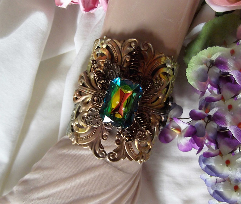 May include: A gold-toned metal cuff bracelet with an ornate design and a large, iridescent green gemstone in the center.
