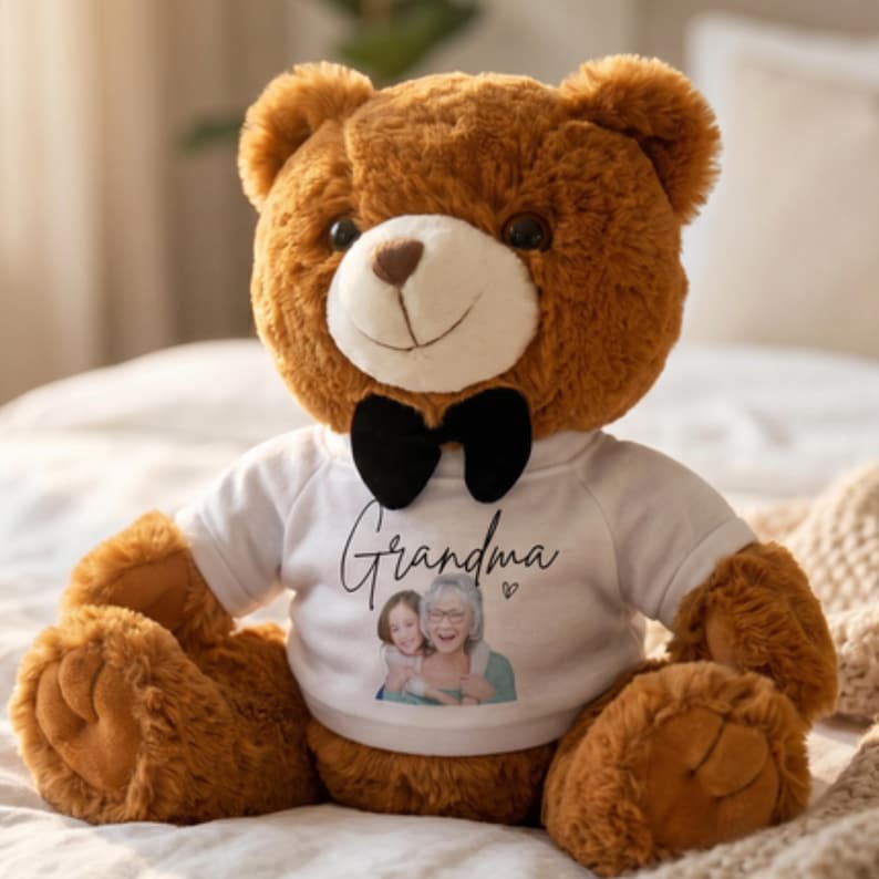 Op de afbeelding: Een bruine teddybeer met een zwarte vlinderdas en een wit t-shirt. Op het t-shirt staat het woord "Grandma" en een foto van een vrouw en een kind. De beer zit op een bed.