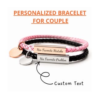 Puede incluir: Dos pulseras personalizadas para parejas. Una es rosa y la otra negra, ambas con correas de cuero trenzado. Cada pulsera tiene una barra de metal grabada con texto y un dije redondo. El texto de las pulseras dice "Her Favorite Mistake" y "His Favorite Problem."