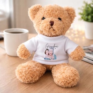 Op de afbeelding: Een lichtbruine teddybeer met een wit t-shirt. Op het shirt staat het woord "Oma" en een afbeelding van een vrouw en een kind. De beer zit op een houten oppervlak, met een witte mok en een plant op de achtergrond.