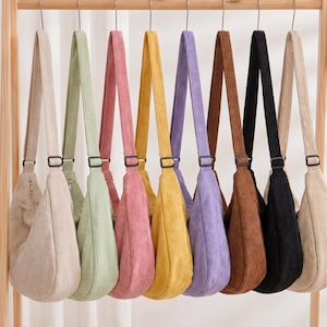 Puede incluir: Una colección de bolsos de hombro de pana en varios colores: beige, verde, rosa, amarillo, morado, marrón, negro y crema. Cada bolso cuelga de una barra de madera, mostrando sus correas ajustables y diseño sencillo.