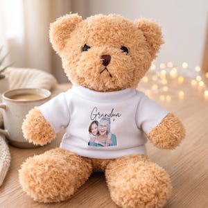 Op de afbeelding: Een bruine teddybeer met een wit t-shirt. Op het shirt staat het woord "Oma" in script boven een portret van een vrouw en een kind. De beer zit op een houten oppervlak.