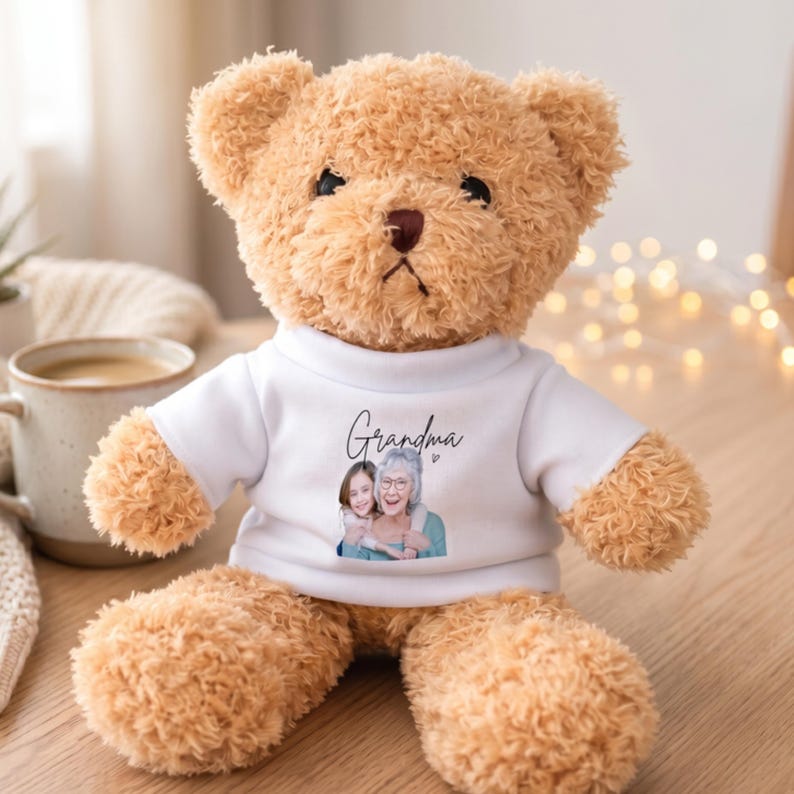 Op de afbeelding: Een bruine teddybeer met een wit t-shirt. Op het shirt staat het woord "Oma" in script boven een portret van een vrouw en een kind. De beer zit op een houten oppervlak.