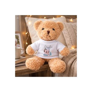 Op de afbeelding: Een zachte, lichtbruine teddybeer met een wit t-shirt. Het t-shirt toont het woord "Oma" in script boven een afbeelding van een vrouw en een meisje. De beer heeft een bruine neus en zit op een houten oppervlak.
